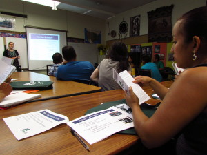 Photo shows a person reading a transit schedule book at a table in a classroom filled with other people at tables facing the front of the room and a screen showing a transit schedule with teacher talking.