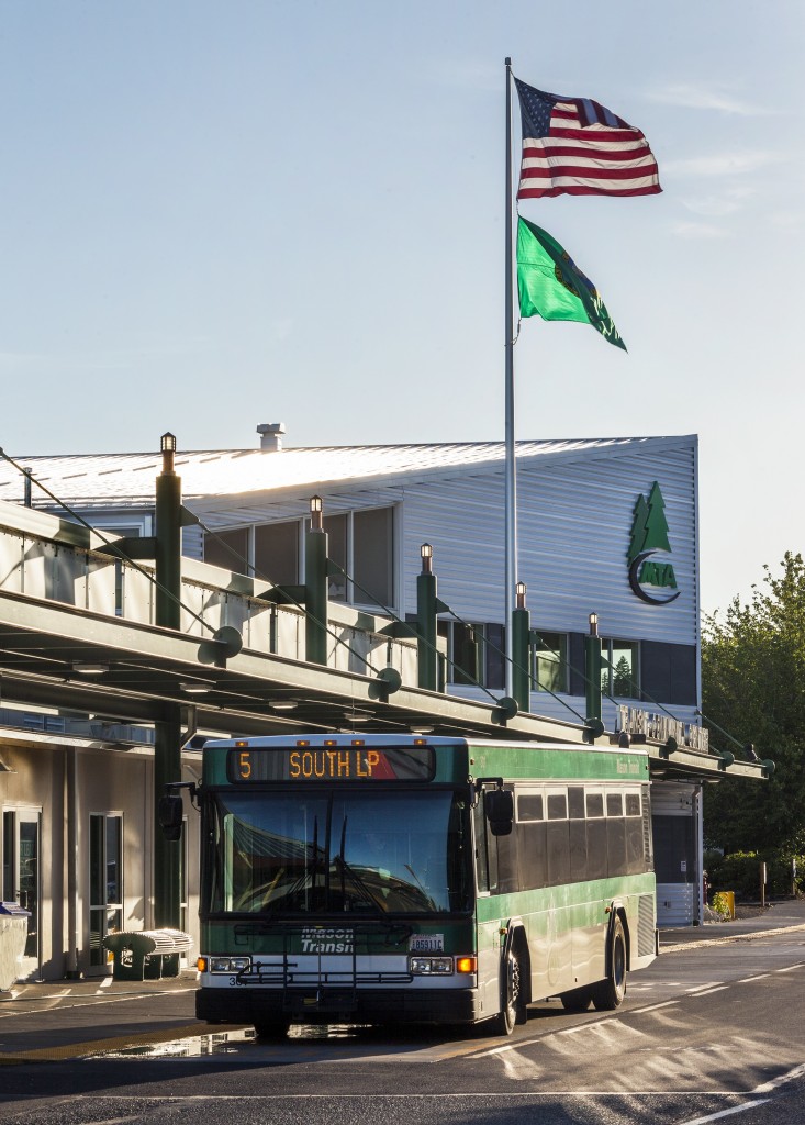 Photo of the front exterior of the Transit-Community Center building at sunset, with the American Flag and State Flag flying and a MTA bus parked in front. 